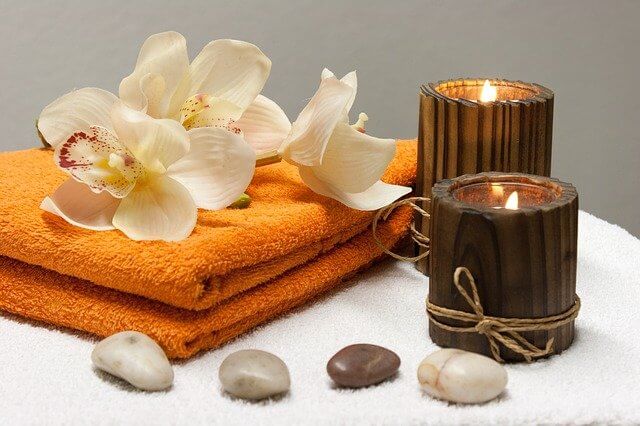 Świece do masażu w brązowych pudełkach obok kamienie do masażu, pomarańczowy ręcznik oraz białe kwiaty